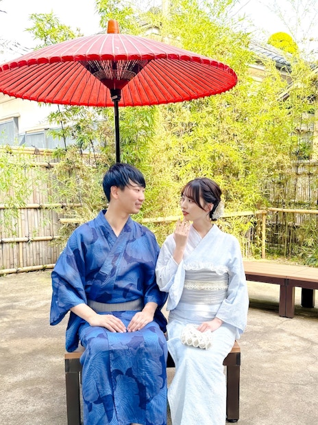 Couple in traditional Japanese kimonos sitting under a red umbrella in a garden.