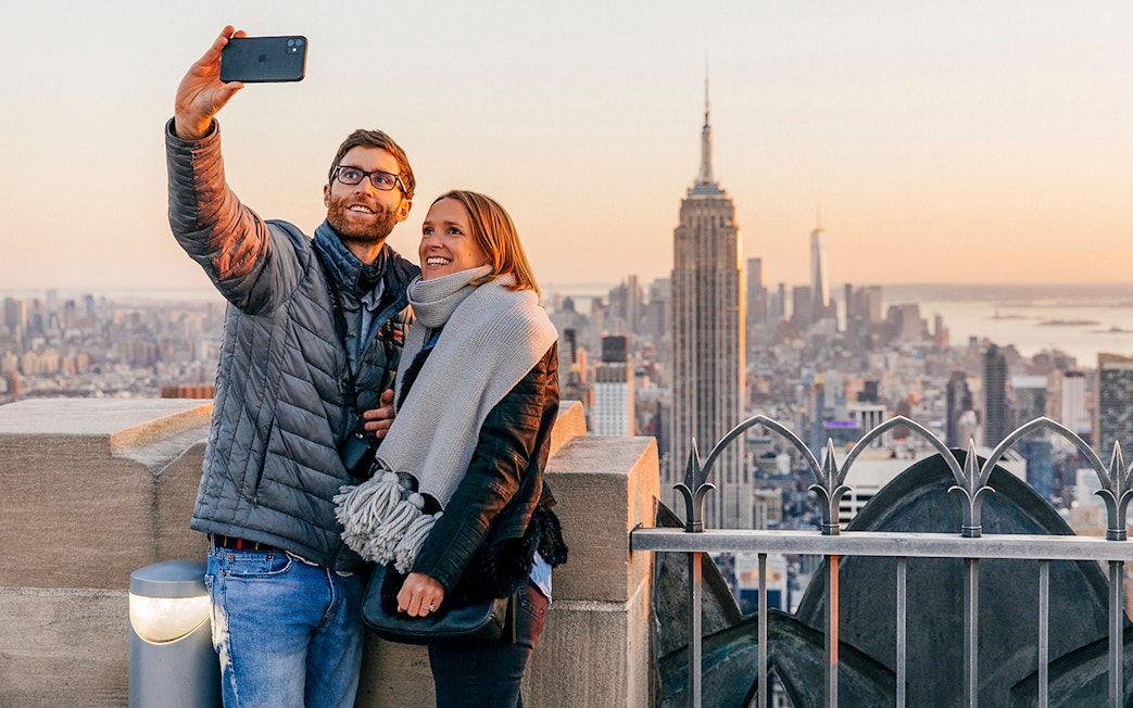 Couple taking selfie at Top of the Rock Observation Deck with Empire State Building in view.