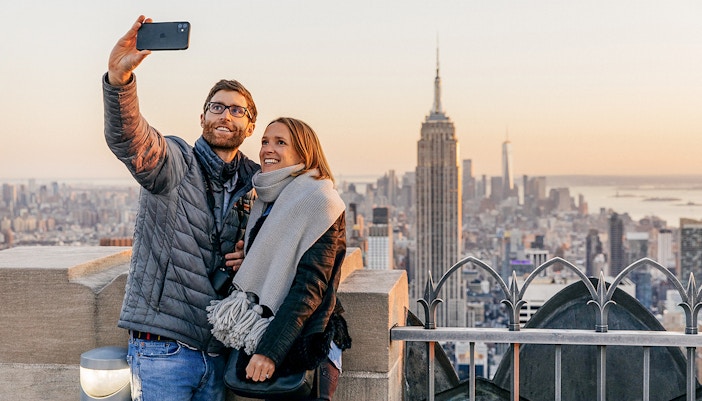 Couple taking selfie at Top of the Rock Observation Deck with Empire State Building in view.