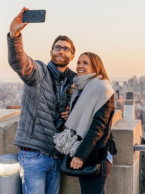 Couple taking selfie at Top of the Rock Observation Deck with Empire State Building in view.