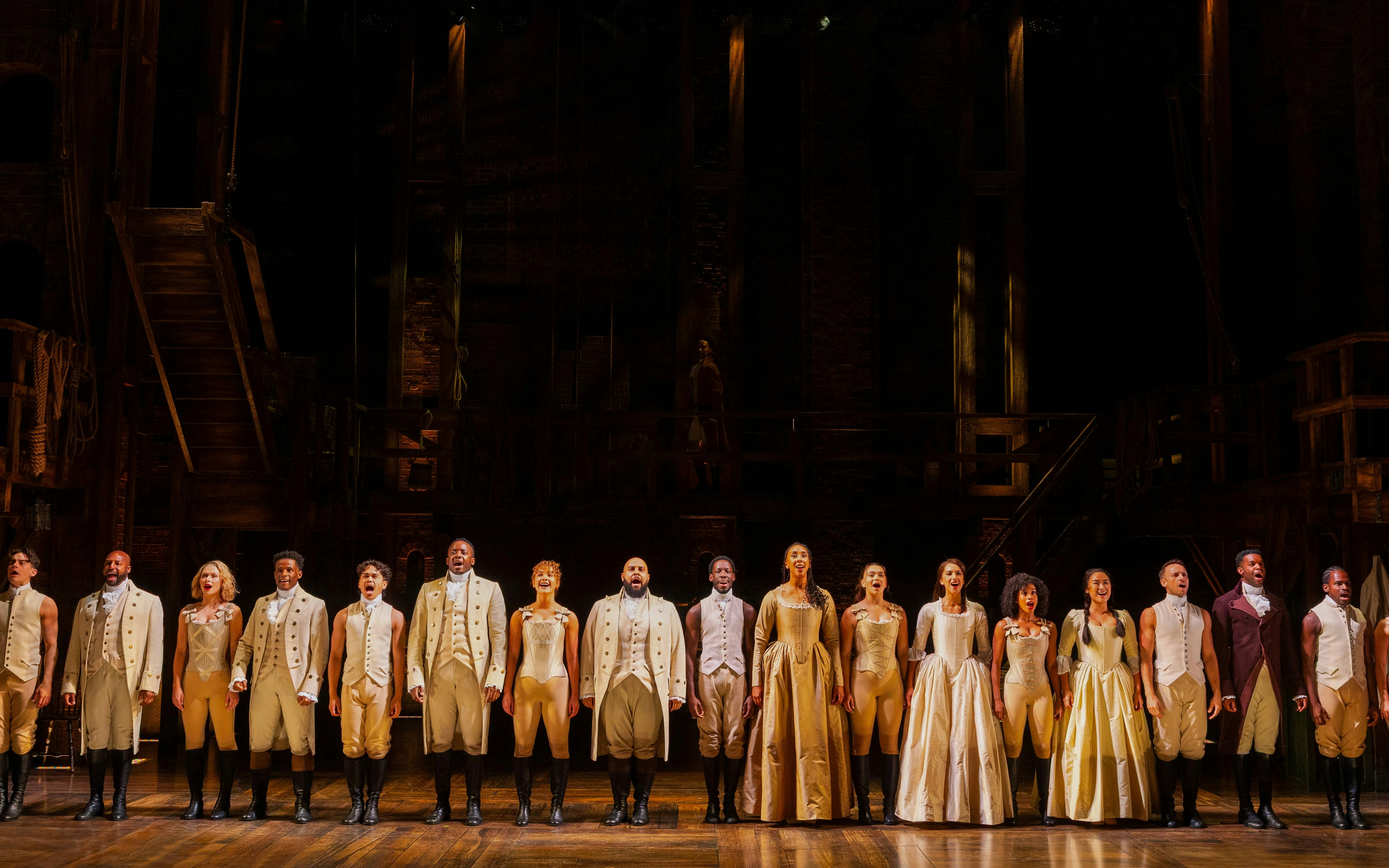 Cast of Hamilton performing on stage in period costumes.