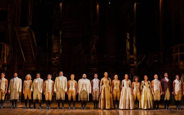 Cast of Hamilton performing on stage in period costumes.