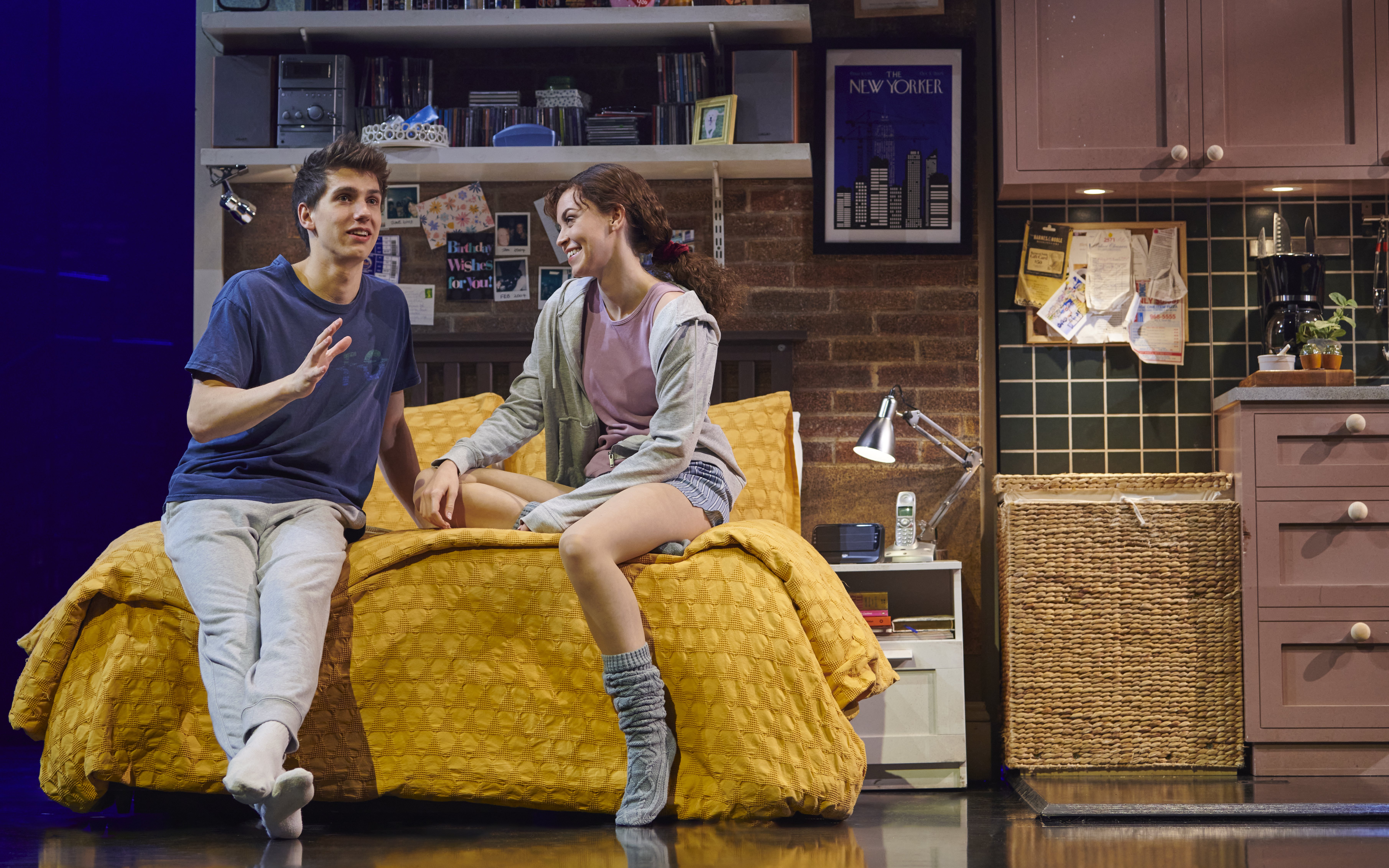 Actors on stage during The Devil Wears Prada West End show, sitting on a bed in a cozy room.