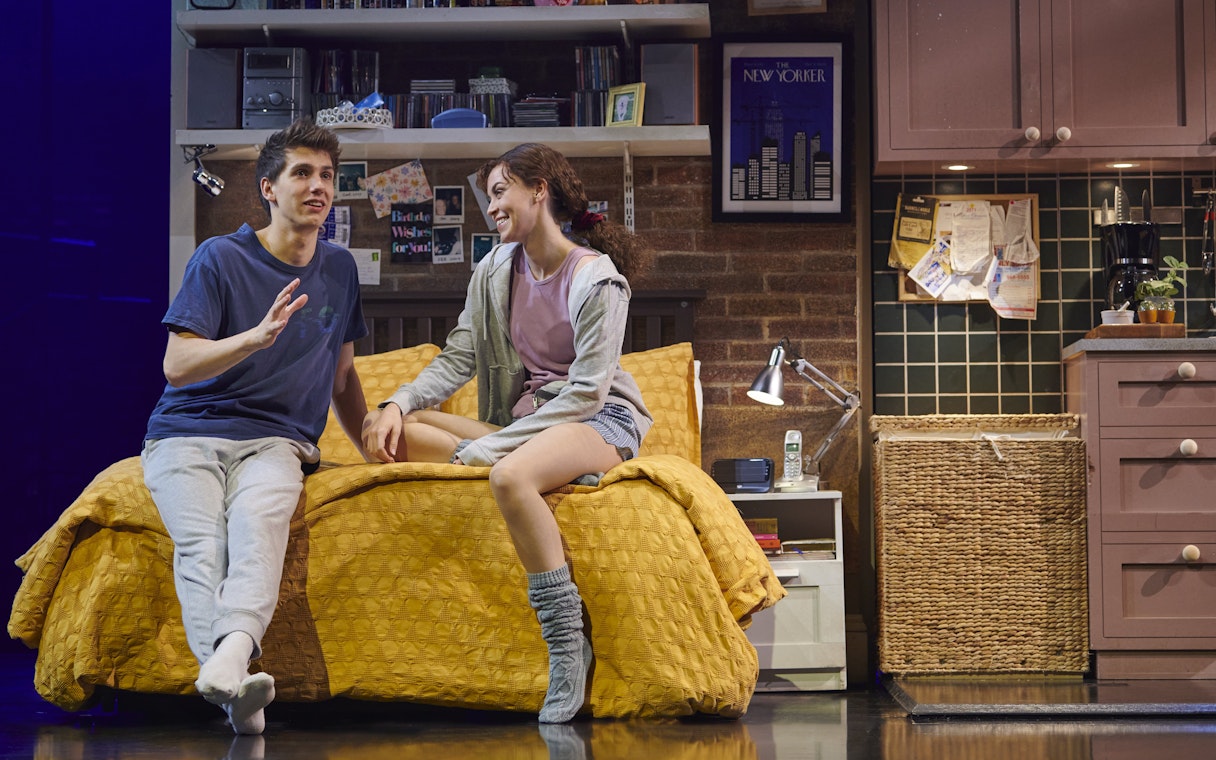 Actors on stage during The Devil Wears Prada West End show, sitting on a bed in a cozy room.