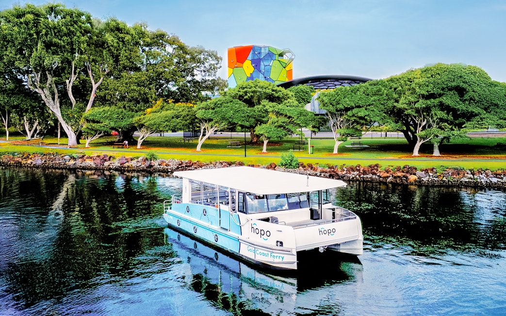 Gold Coast ferry near Home of the Arts with colorful building and lush trees in the background.