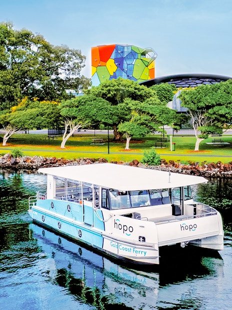 Gold Coast ferry near Home of the Arts with colorful building and lush trees in the background.