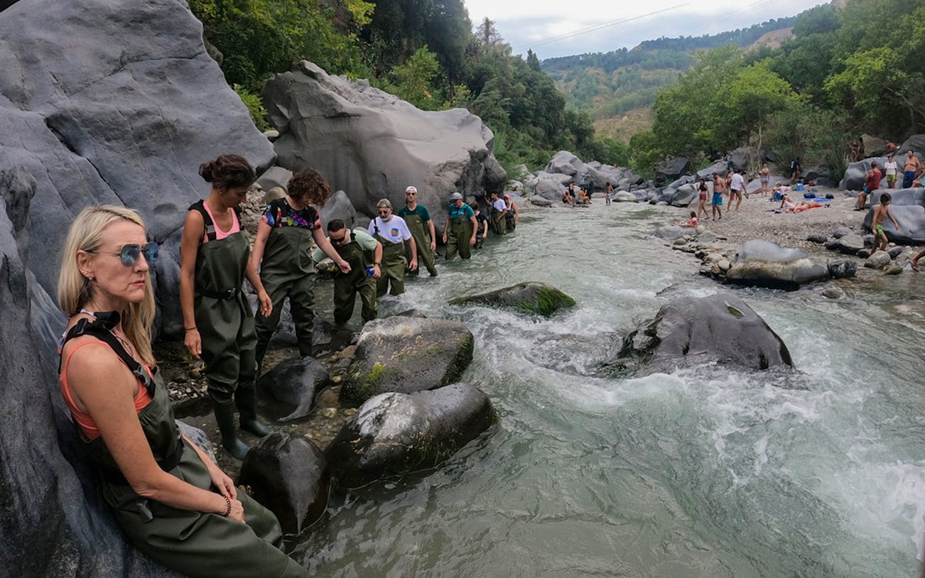 Group river trekking in Gole Alcantara, Sicily, wearing waders and exploring rocky terrain.