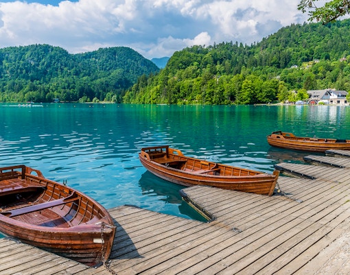 Boats moored along the shore of Lake Bled with forested hills in the background.
