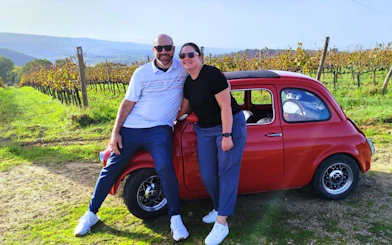 Couple leaning on red vintage Fiat 500 in Tuscany vineyard.