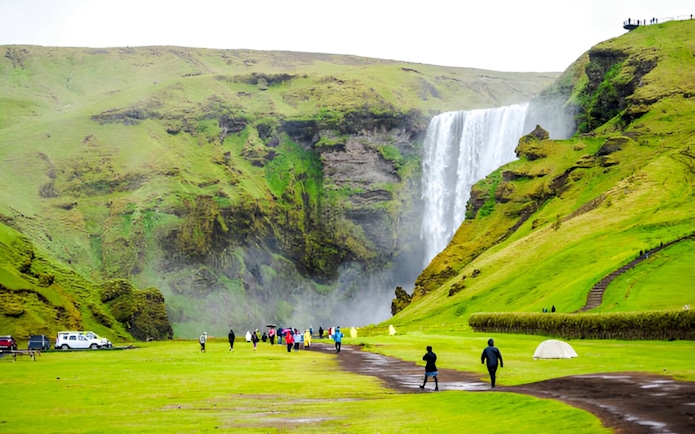 Visitors walking towards Skógafoss waterfall in Iceland, surrounded by lush green hills.