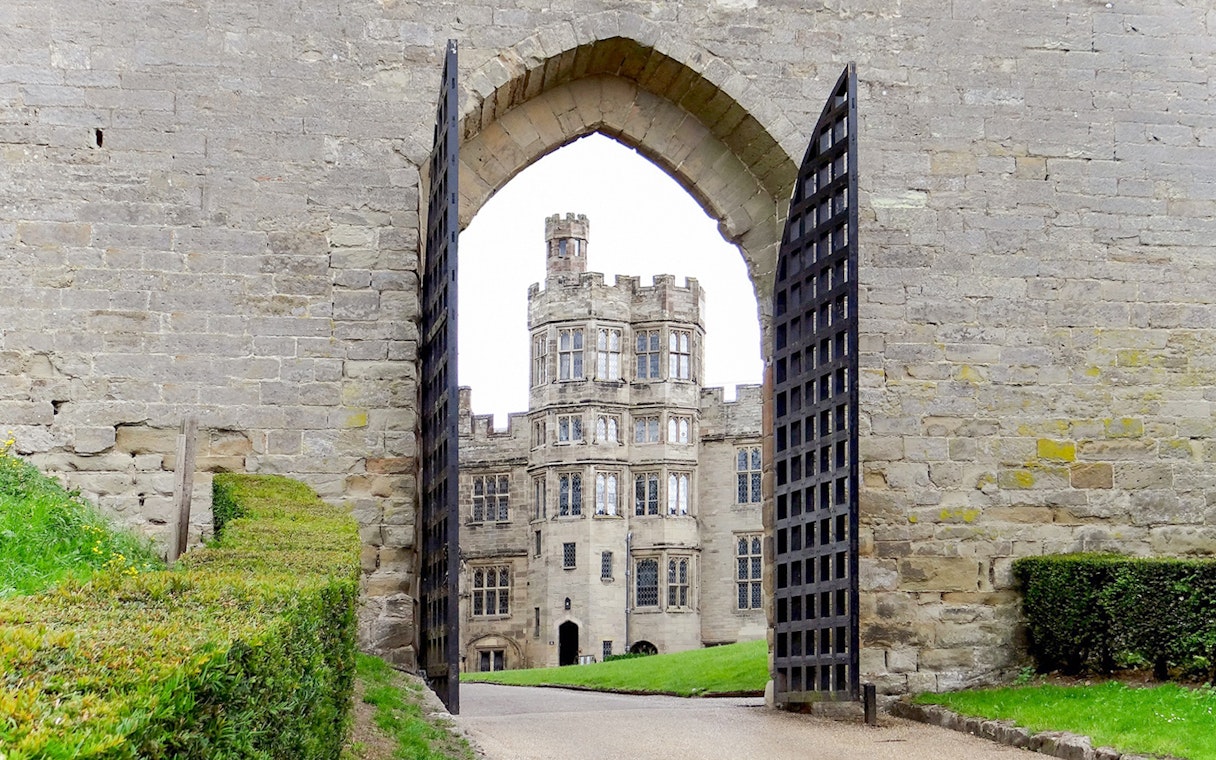 Warwick Castle gate open with view of historic stone building and manicured hedges.