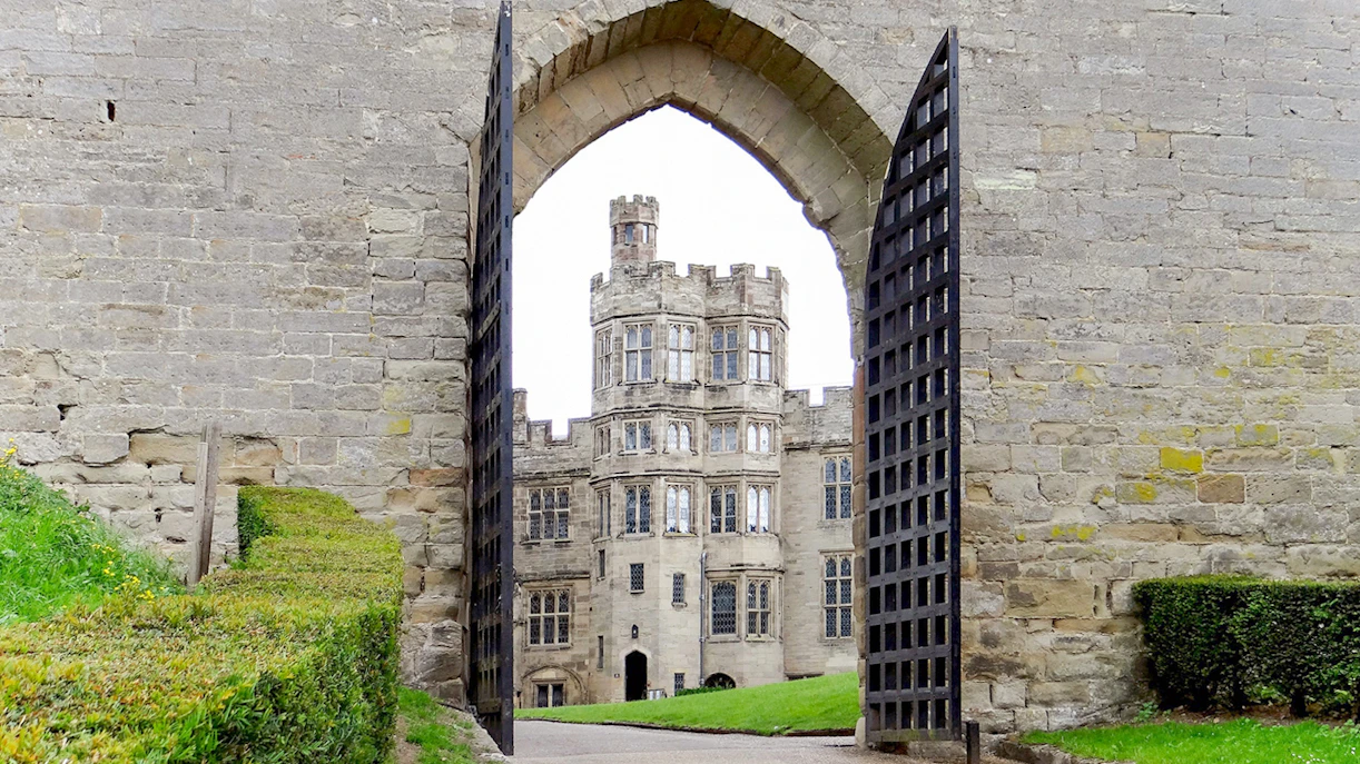 Warwick Castle gate open with view of historic stone building and manicured hedges.