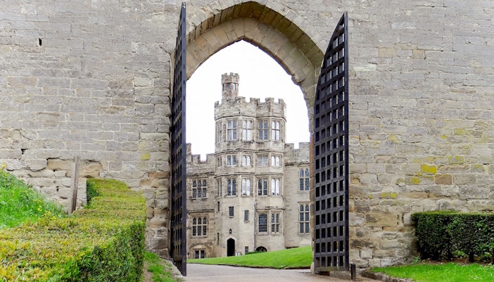 Warwick Castle gate open with view of historic stone building and manicured hedges.