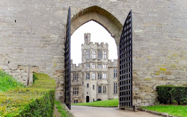 Warwick Castle gate open with view of historic stone building and manicured hedges.
