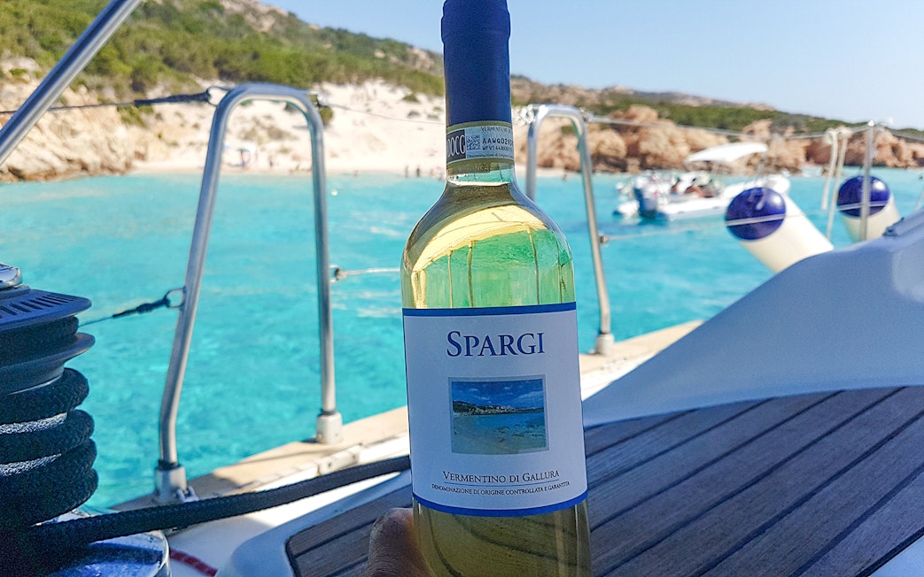 Sailboat deck with wine bottle, turquoise waters of La Maddalena Archipelago in background.