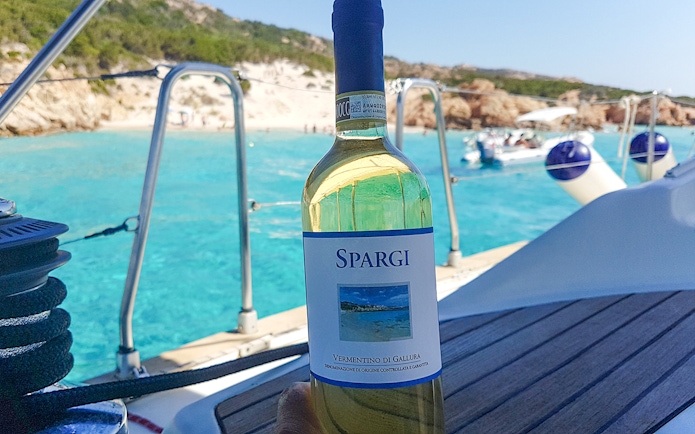 Sailboat deck with wine bottle, turquoise waters of La Maddalena Archipelago in background.