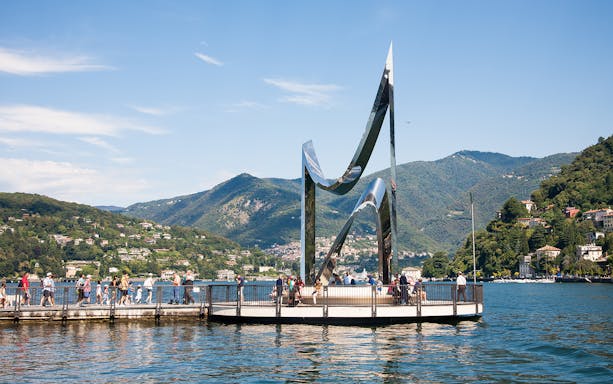 People walking on a pier near the Life Electric sculpture on Lake Como, Italy, with mountains in the background.