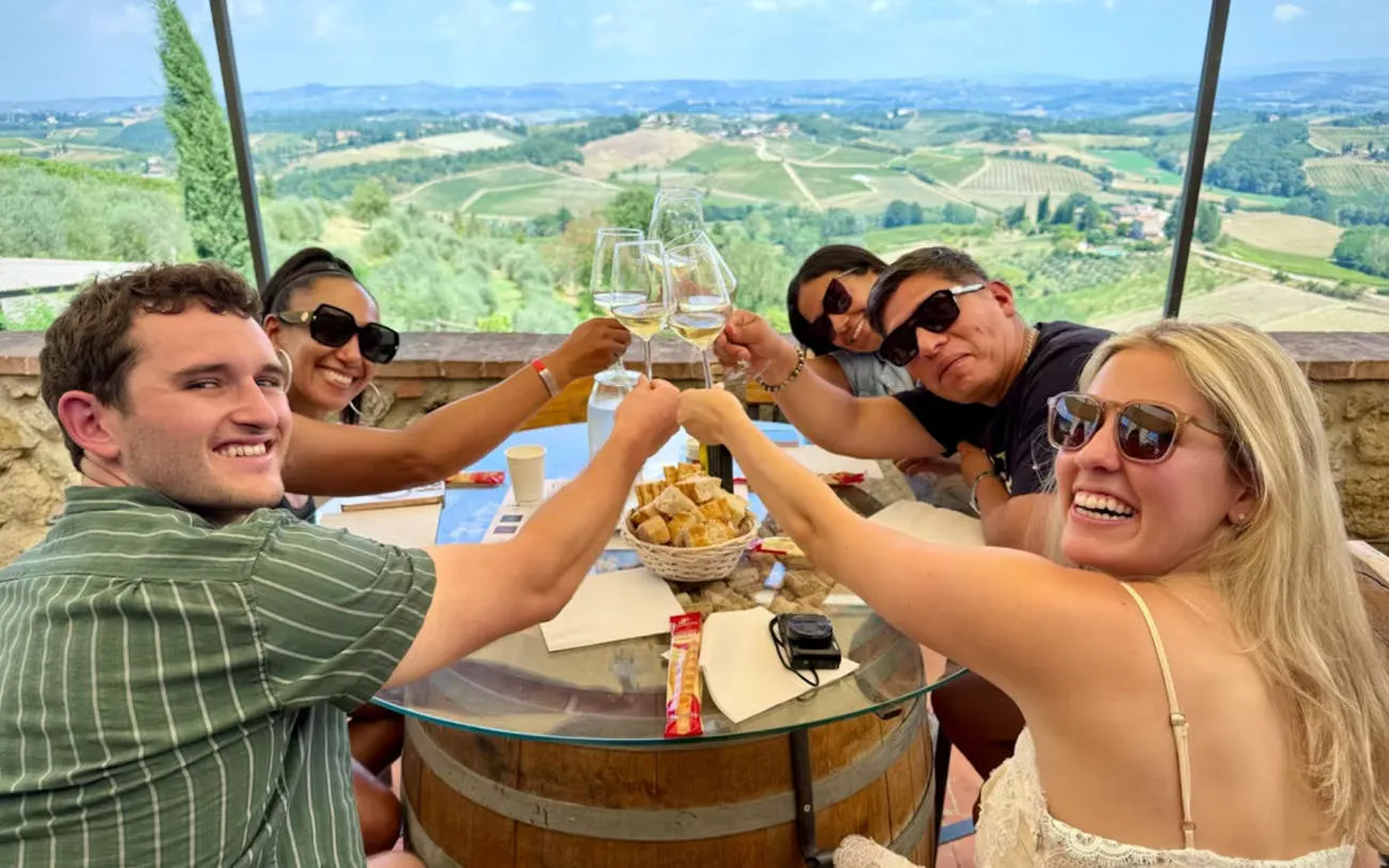Tourists enjoying wine tasting at a table overlooking Tuscany vineyards.
