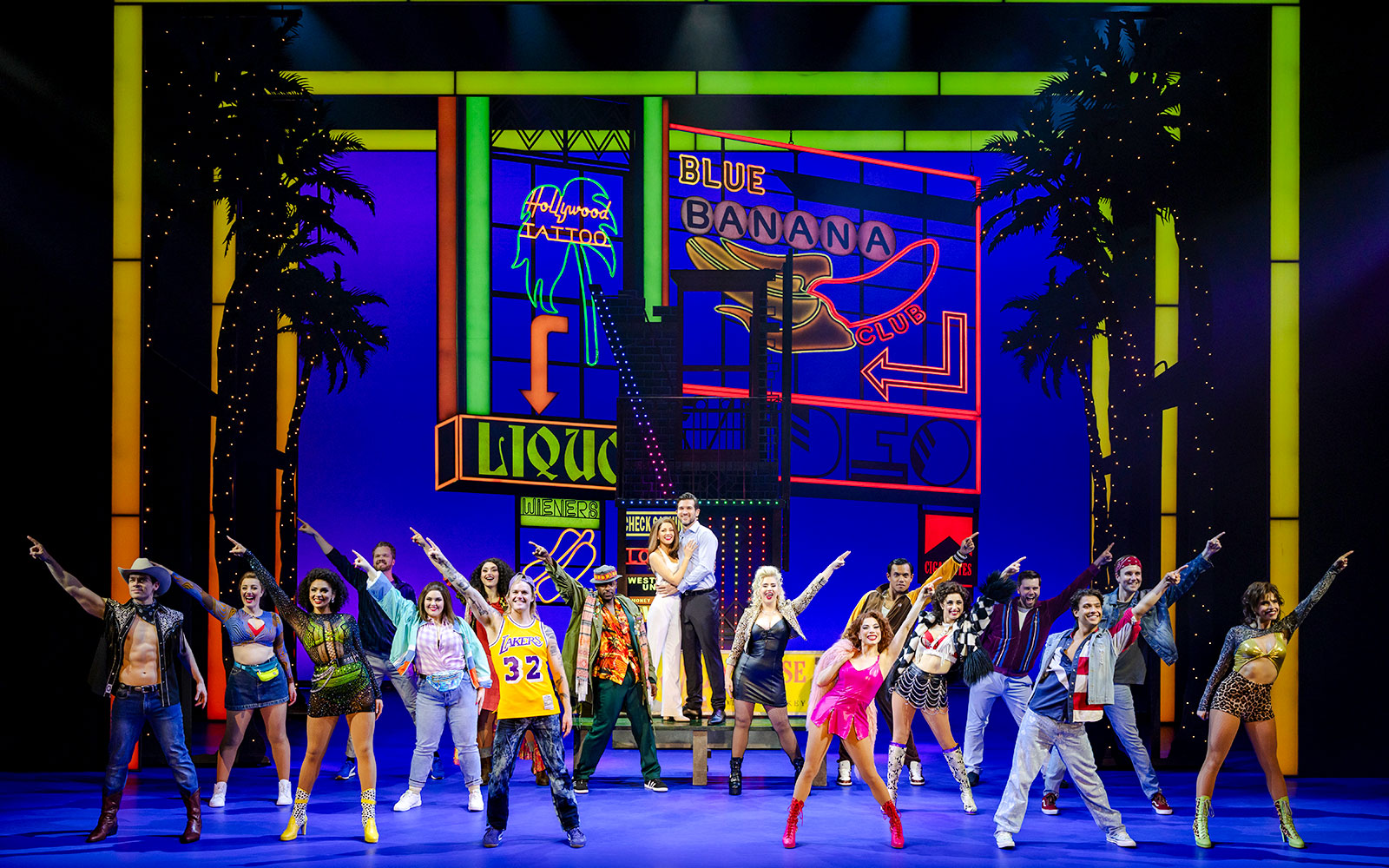 Cast performing a scene from Pretty Woman The Musical with colorful neon signs in the background.