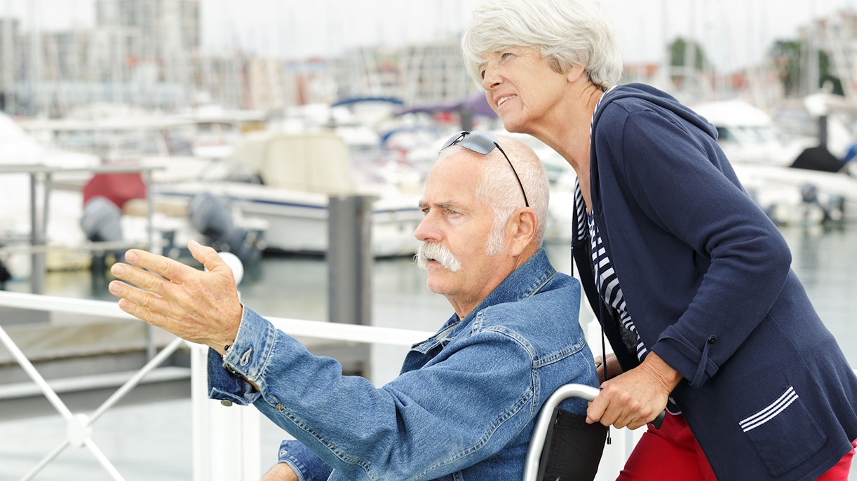 Elderly man in wheelchair at a marina, pointing towards boats.