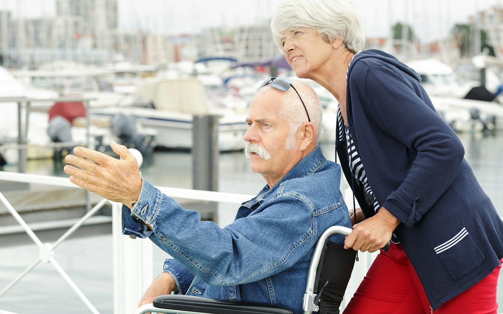 Elderly man in wheelchair at a marina, pointing towards boats.