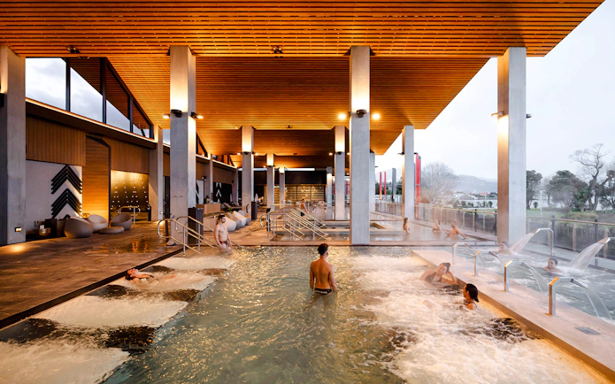 Visitors enjoying thermal pools at Wai Ariki Hot Springs & Spa, with modern architecture and scenic views.