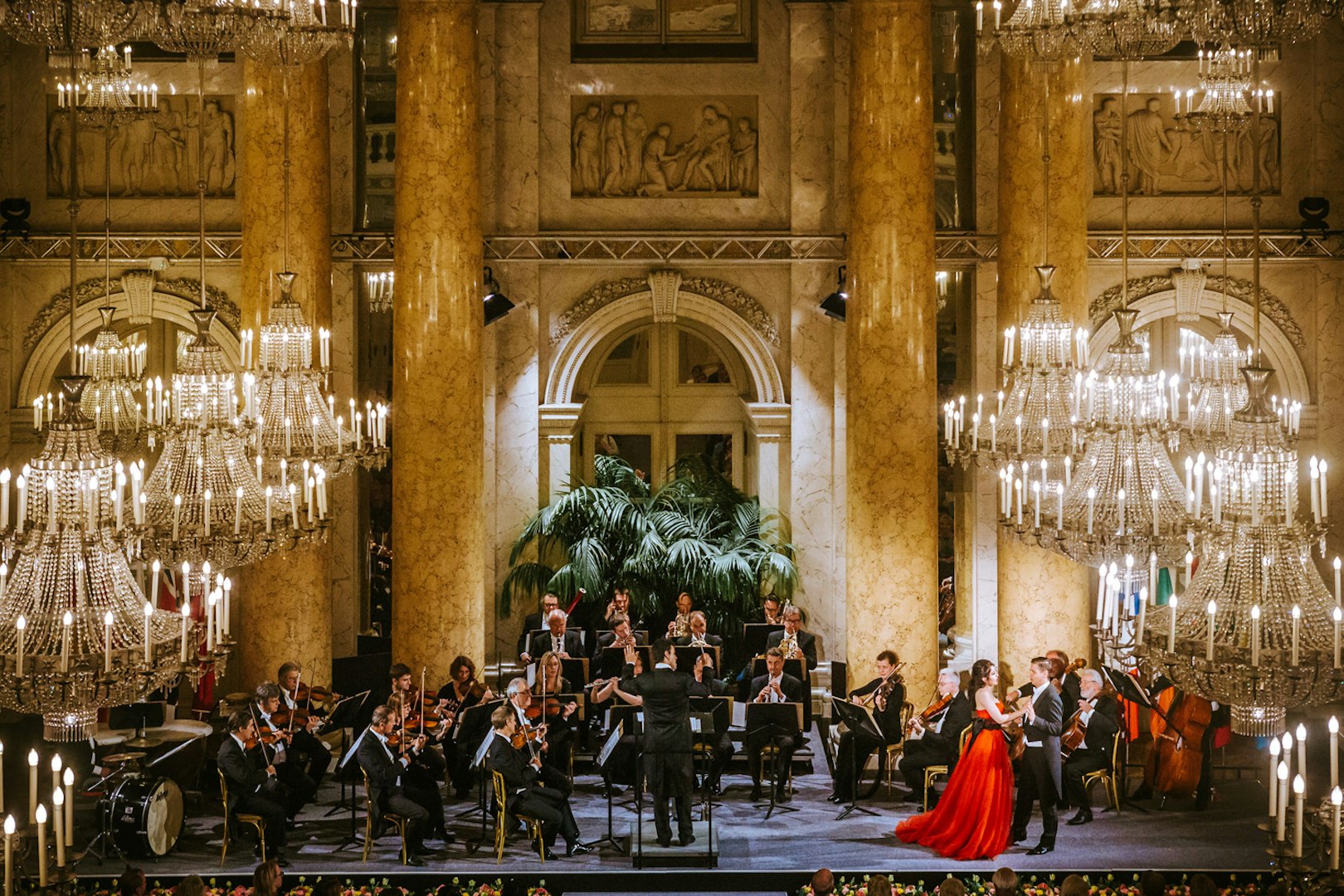 Vienna Hofburg Orchestra performing in an ornate hall with chandeliers.