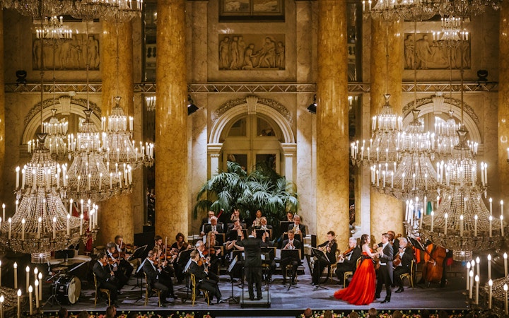 Vienna Hofburg Orchestra performing in an ornate hall with chandeliers.