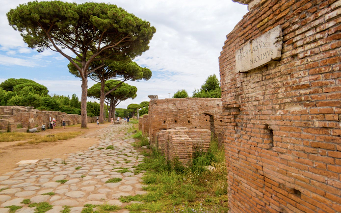 Ancient stone road and ruins in Ostia Antica, Italy, with pine trees lining the path.
