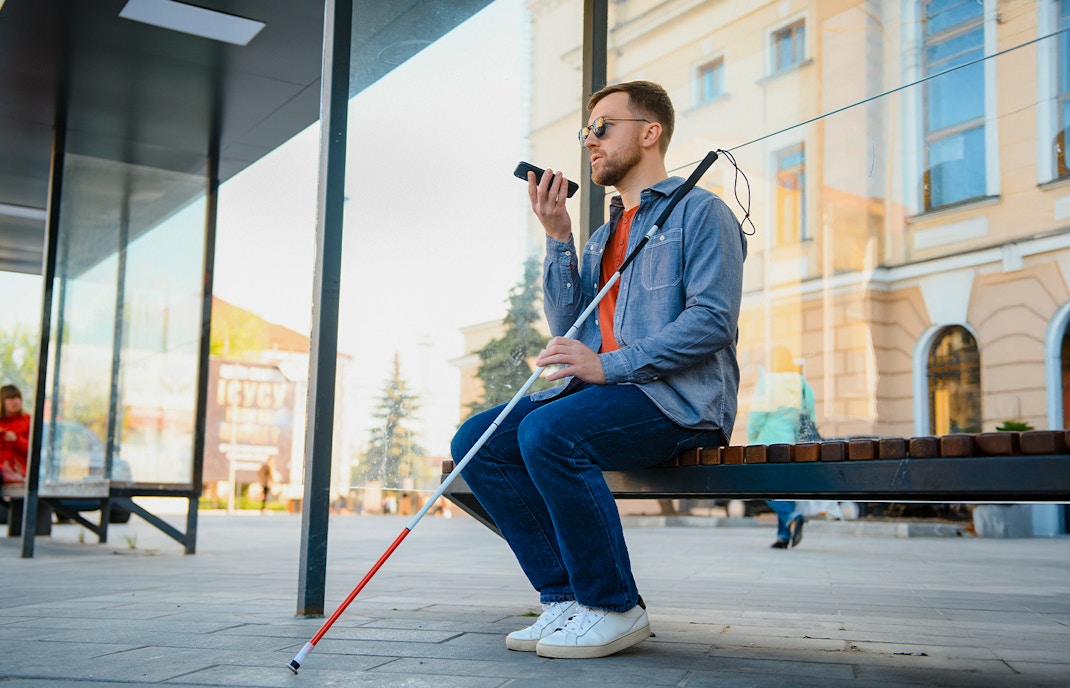 Blind man with walking stick and glasses sitting on a bench outside a museum.