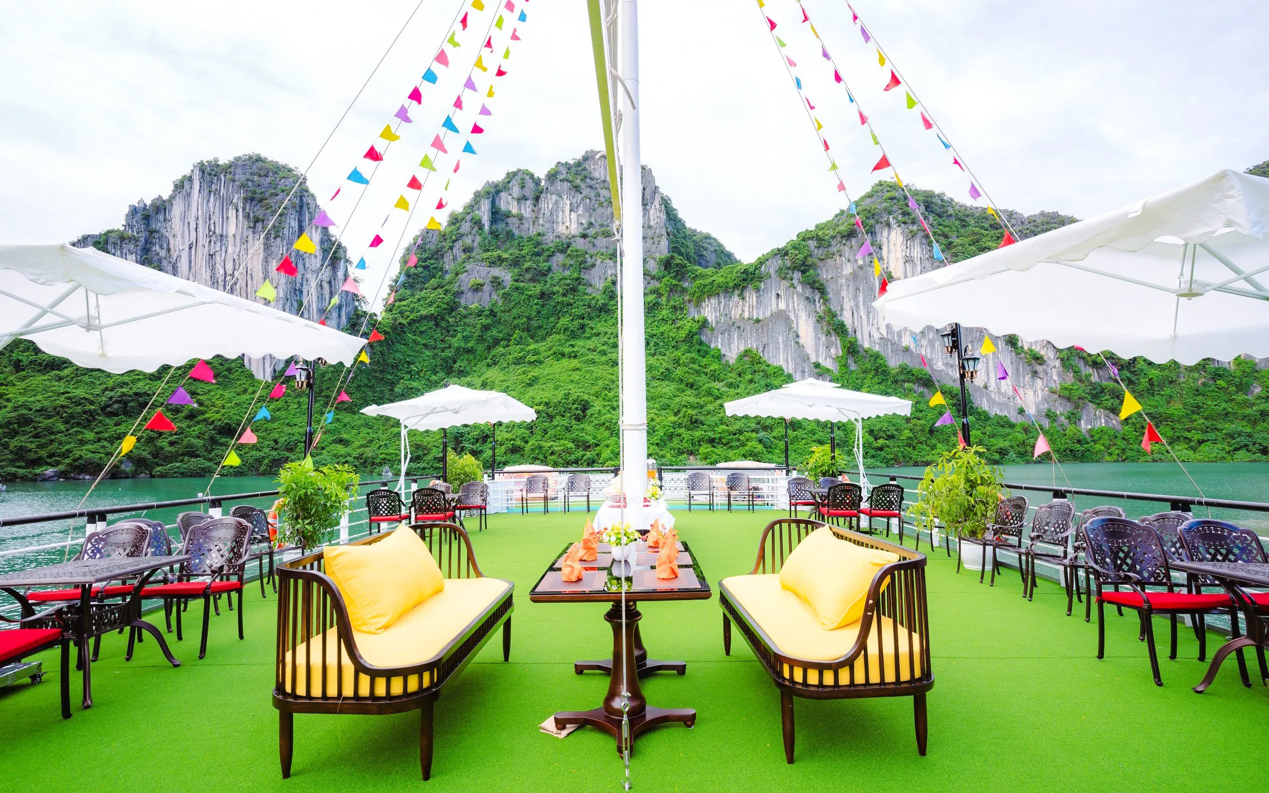 Deck of Apollo Cruise with seating and colorful flags, Ha Long Bay limestone cliffs in background.