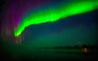 Northern Lights over a lake near Rovaniemi, Finland, with a forest silhouette.
