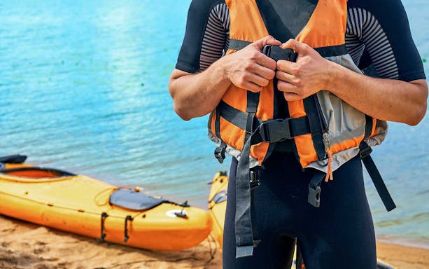 Kayaker fastening life vest near orange kayak on sandy shore.