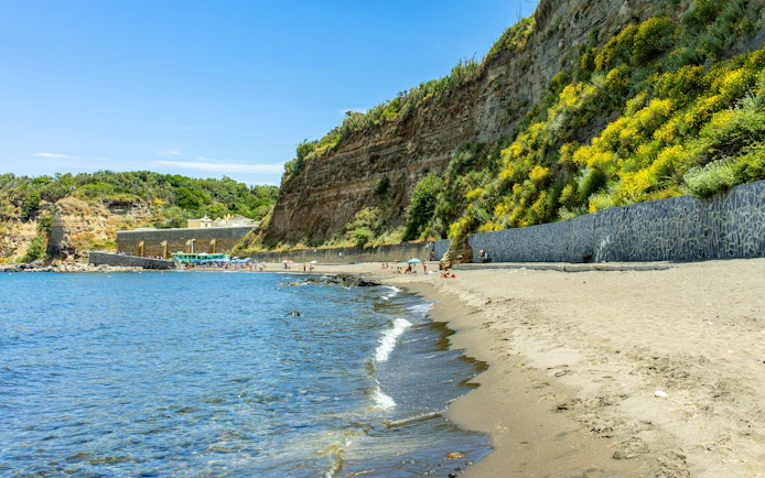 Sandy shoreline and cliffs at Postino Beach with people relaxing by the water.