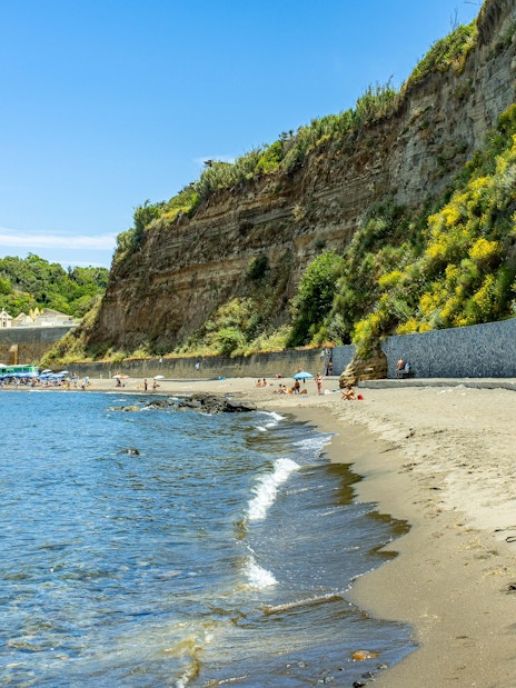Sandy shoreline and cliffs at Postino Beach with people relaxing by the water.