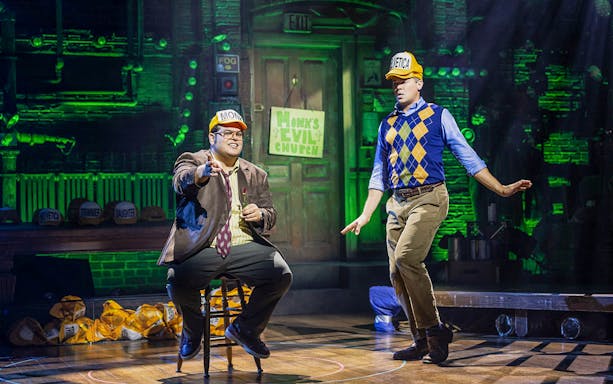Two actors performing on stage in "Gutenberg! The Musical!" with hats labeled Monk and Helvetica.