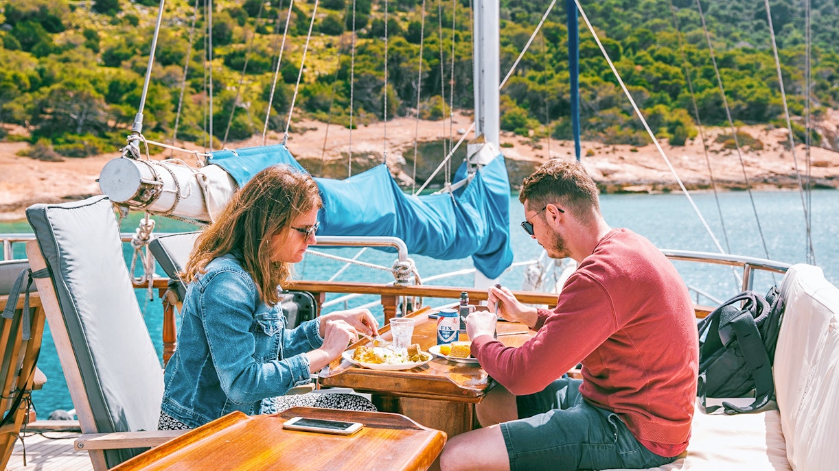 Couple enjoying lunch with beer on a cruise ship deck near a scenic coastline.