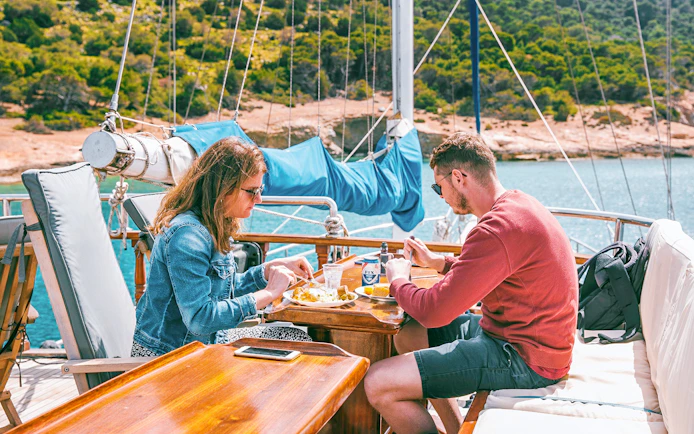 Couple enjoying lunch with beer on a cruise ship deck near a scenic coastline.