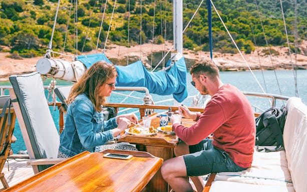 Couple enjoying lunch with beer on a cruise ship deck near a scenic coastline.