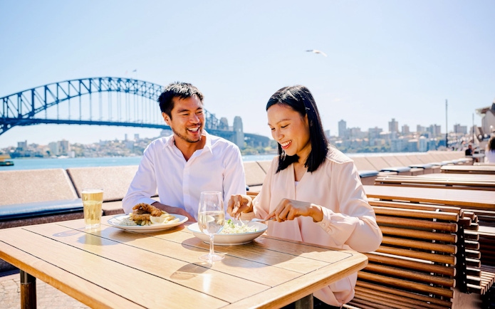 Couple dining outdoors with Sydney Harbour Bridge in the background.