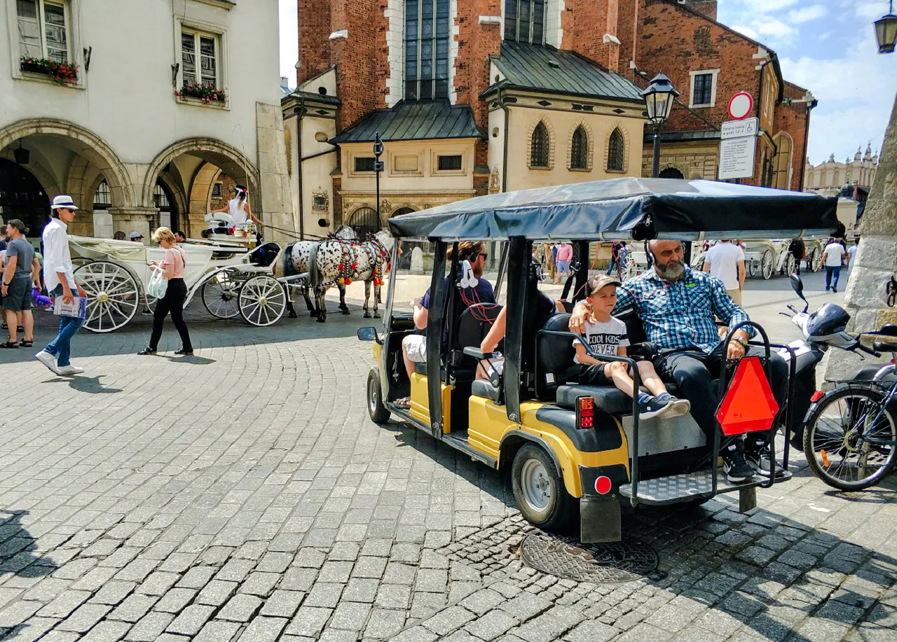 Golf cart tour in Krakow's Old Town with horse-drawn carriages and historic buildings in the background.