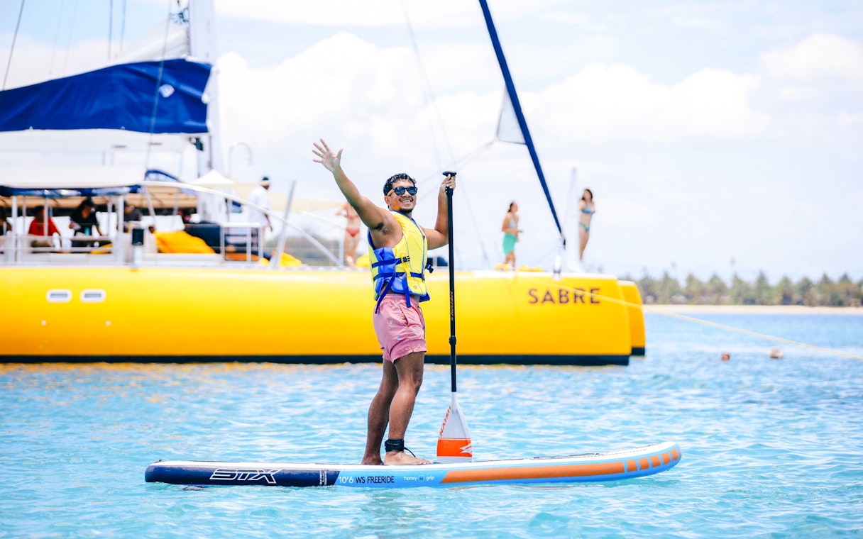 Person paddle surfing near a yellow catamaran, South Sea Sailing, Fiji.