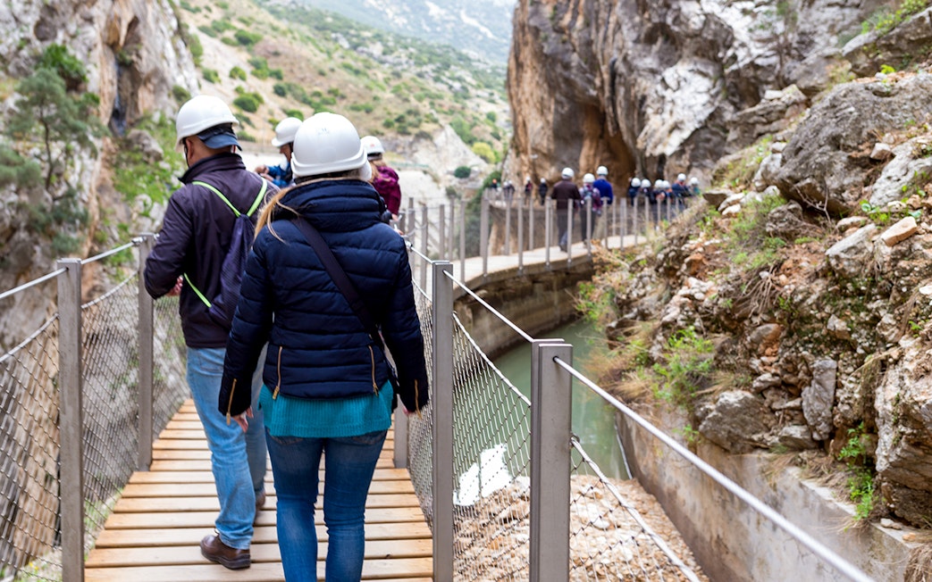 Visitors walking on Caminito del Rey path with helmets, surrounded by cliffs and river in Spain.