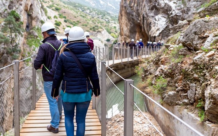 Visitors walking on Caminito del Rey path with helmets, surrounded by cliffs and river in Spain.
