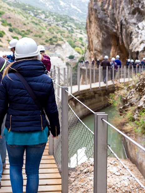 Visitors walking on Caminito del Rey path with helmets, surrounded by cliffs and river in Spain.