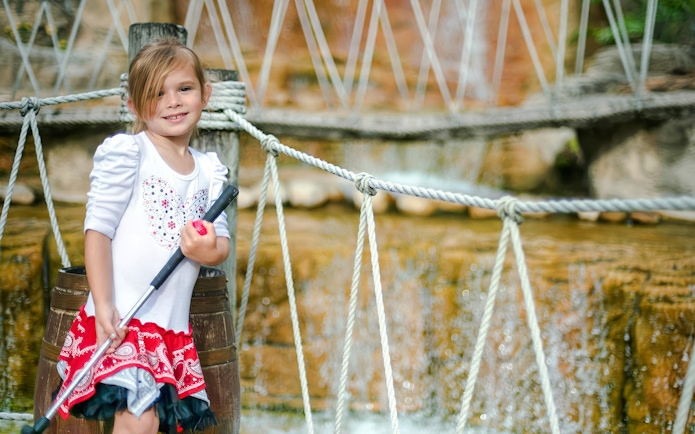 Child holding a mini-golf club at Pirate's Cove Adventure Golf with rope bridge in background.