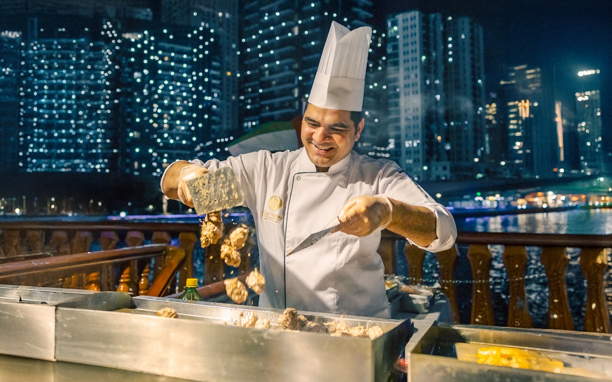 Chef grilling meat on Alexandra VIP Dhow Cruise with city skyline in background.