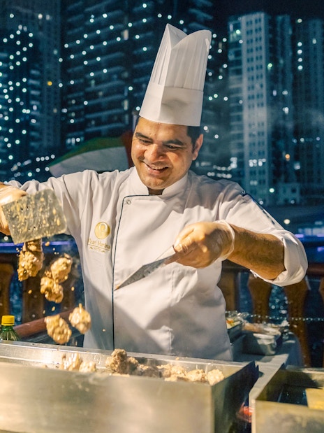 Chef grilling meat on Alexandra VIP Dhow Cruise with city skyline in background.