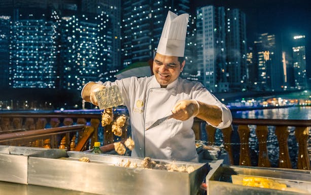 Chef grilling meat on Alexandra VIP Dhow Cruise with city skyline in background.