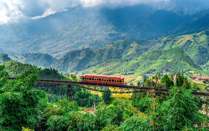 Red train crossing a bridge with Fansipan mountain landscape at Sun World Fansipan Legend, Vietnam.
